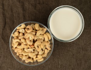 A glass of cashew milk with a bowl of cashews on a brown tablecloth.