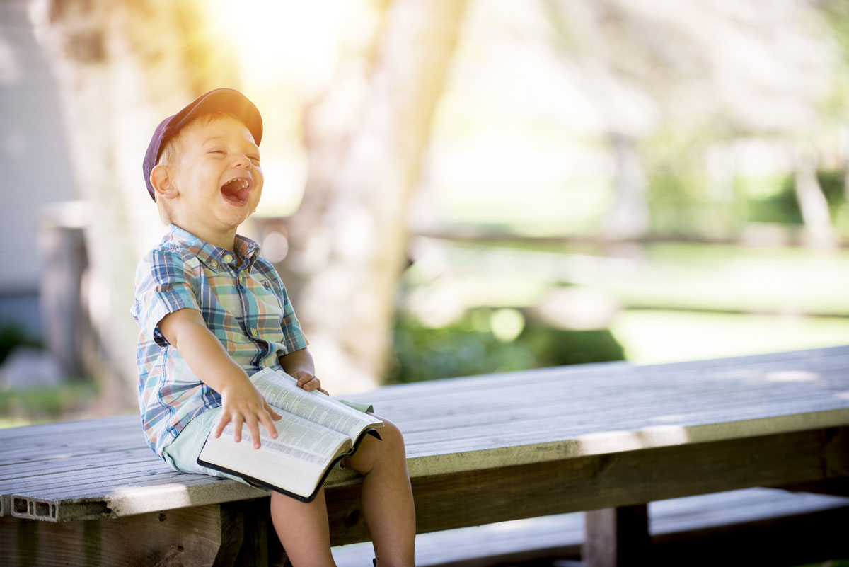 little boy sitting on a bench, holding a book, and laughing