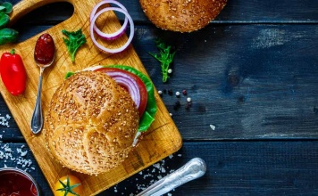 cutting board with a burger and a hearty bun with onion, pepper, and ketchup on the side