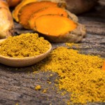 the turmeric powder in spoon and roots on wooden plate
