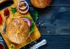 cutting board with a burger and a hearty bun with onion, pepper, and ketchup on the side